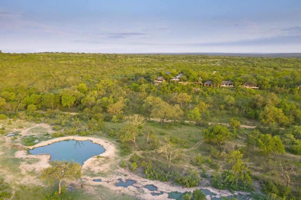 Aerial view of a savannah with green trees and a cluster of thatched-roof lodges in the distance, beside a irregular blue pond in the foreground.