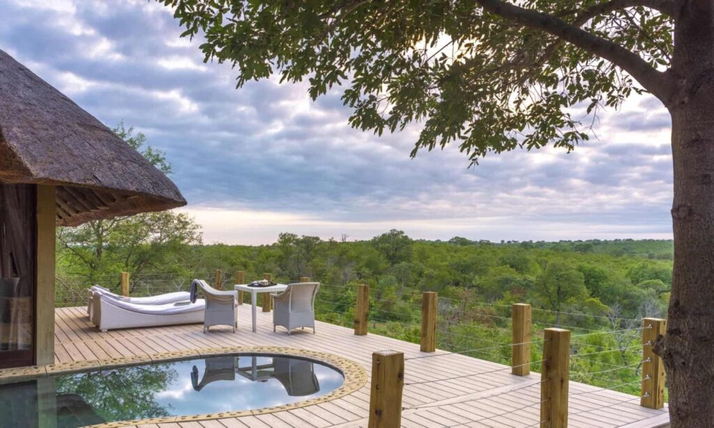 Wooden deck with a circular pool, white lounge chairs, and a small table, overlooking a green savanna under a cloudy sky.