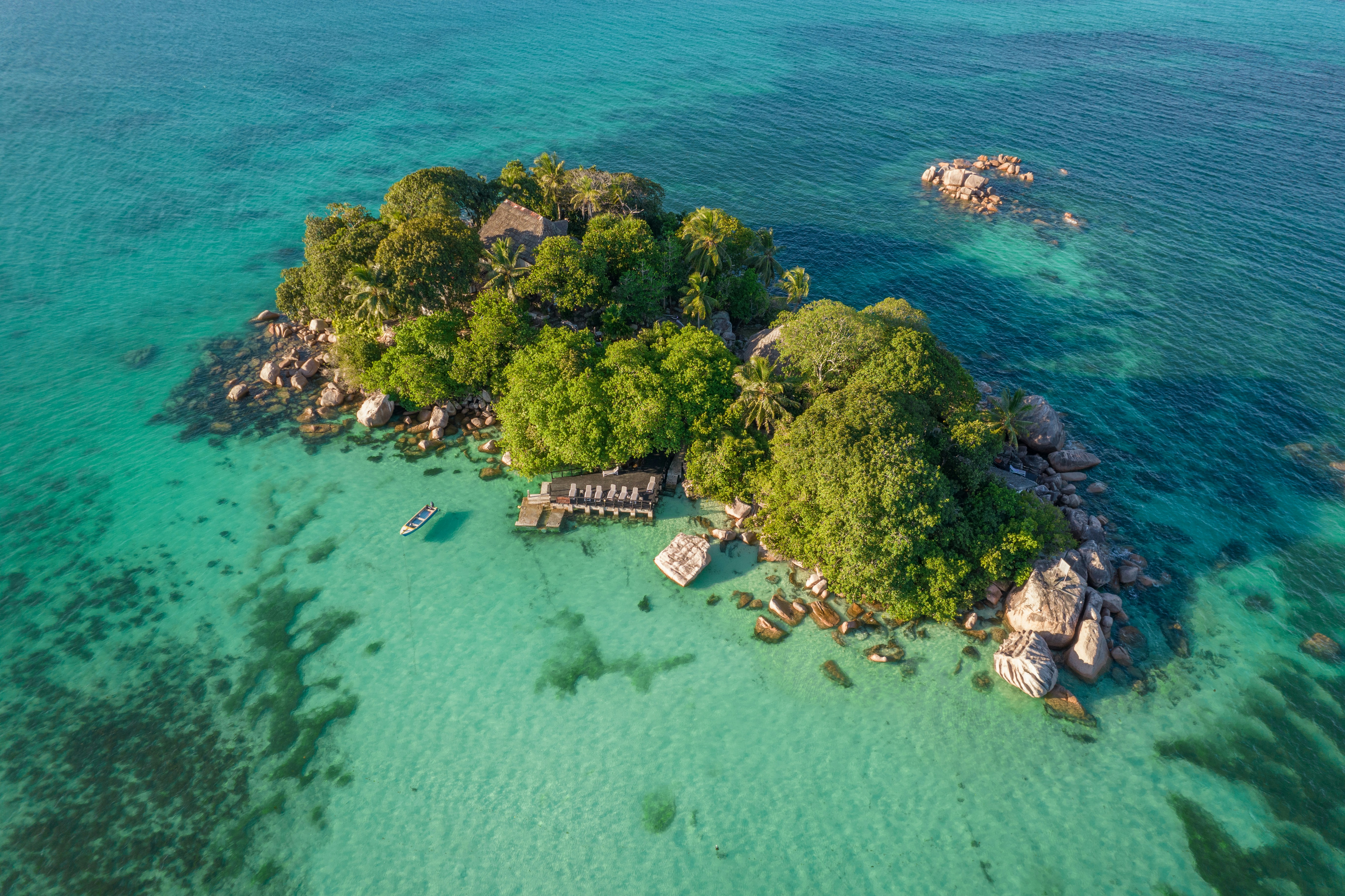 A small tropical island with dense green trees, rocky shore, and turquoise shallow water viewed from above.