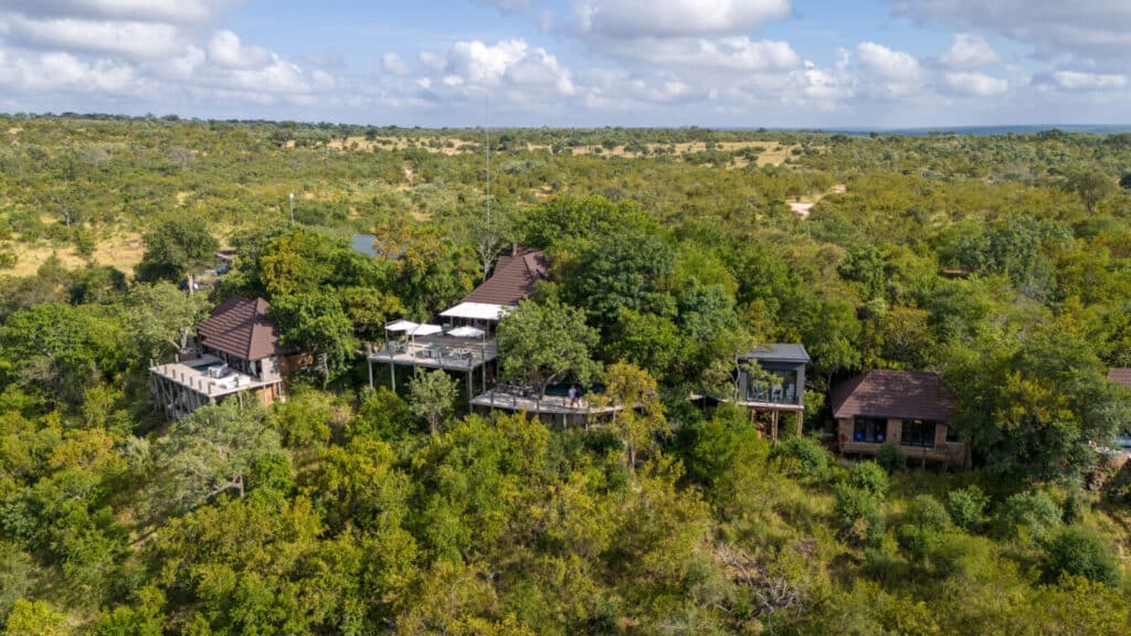 Aerial view of a wooden lodge complex on stilts nestled among dense green trees in a savannah landscape, with terraces and umbrellas on decks interspersed by buildings on slopes.