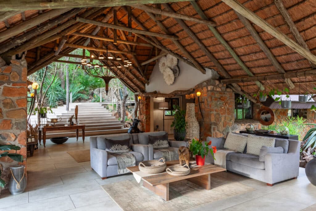Rustic living room with exposed timber roof, stone walls, and gray sectional sofas around a wooden coffee table with plants and a chandelier overhead. A staircase and greenery are visible at the back.