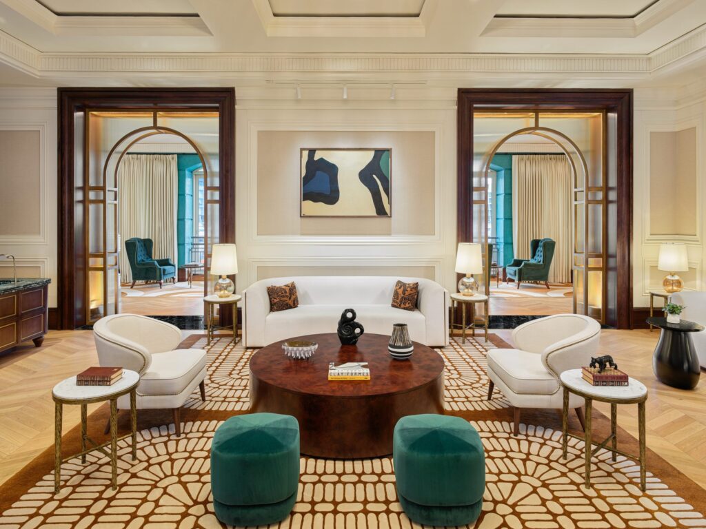 Luxurious hotel lounge with cream walls, a white sofa, teal ottomans, and a dark wood round coffee table in a patterned rug setting with arched doorways nearby.