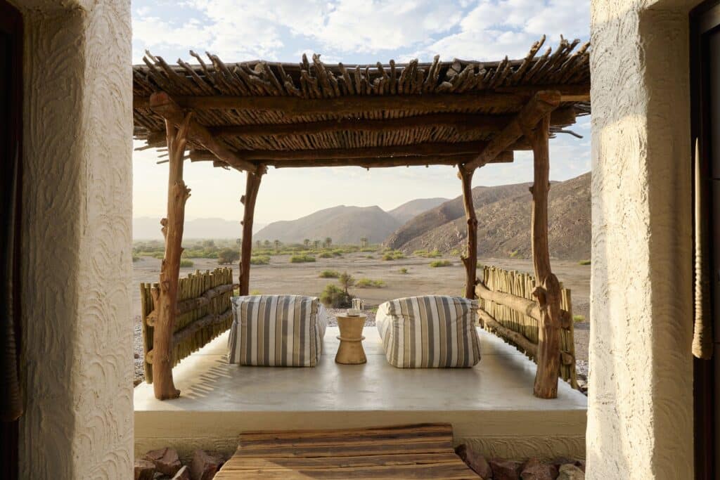 Open-air seating area under a rustic wooden pergola with striped cushions, desert landscape beyond.