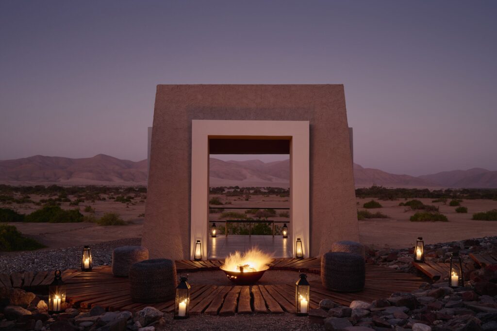 Desert scene at dusk with a large rectangular stone archway, lantern-lit path, and a central fire pit on a wooden deck.