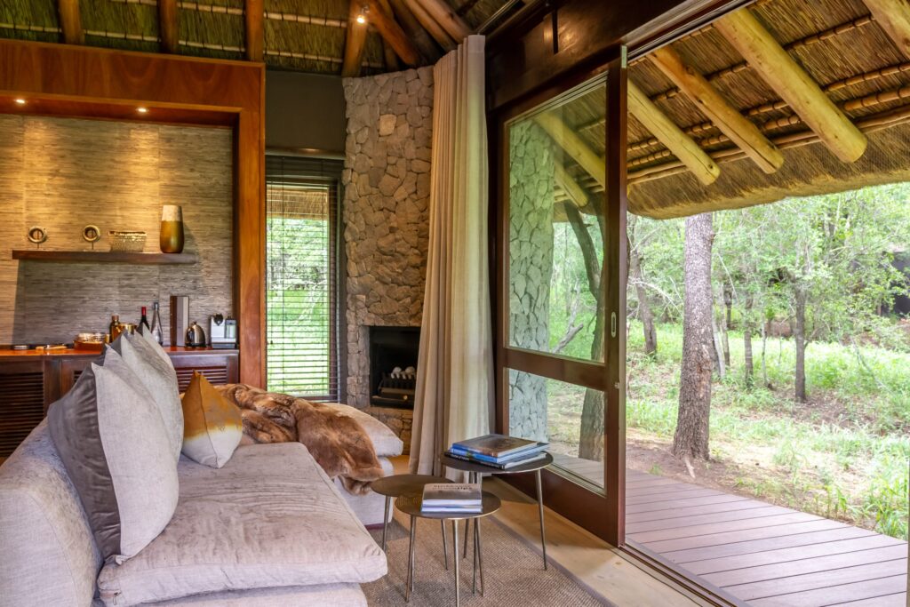 Cozy living room with a beige sectional sofa, cushions, and a fur throw, plus a stone wall and wooden beams. A glass door opens to a wooded outdoor view.