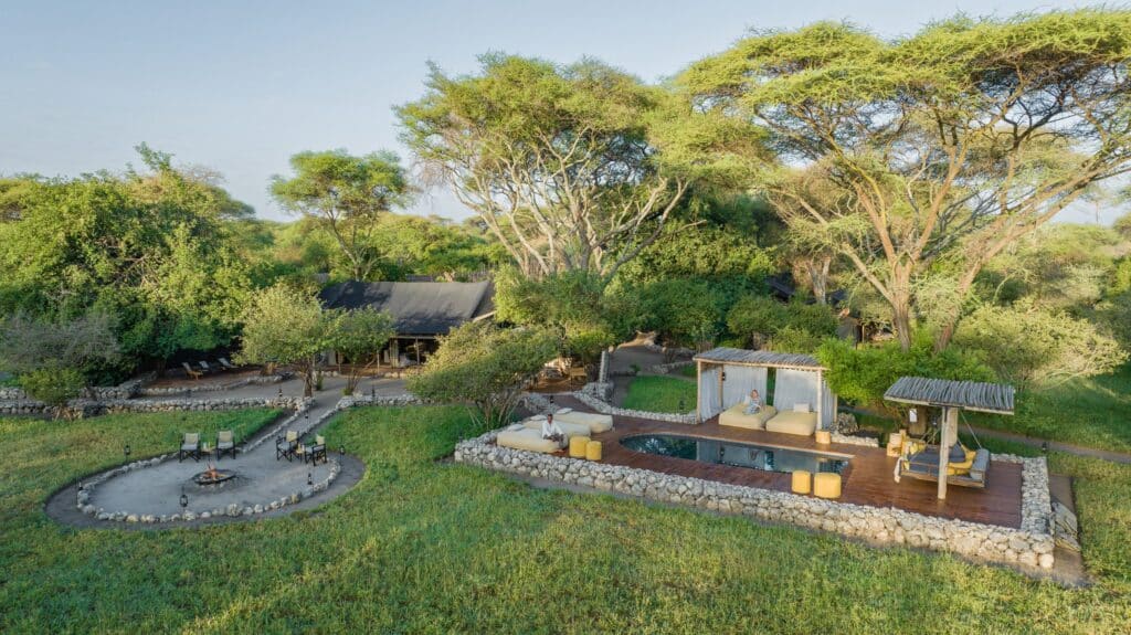 Aerial view of a green resort yard with stone terraces, circular fire pit, lounge seating, cabanas, and a pool among trees.