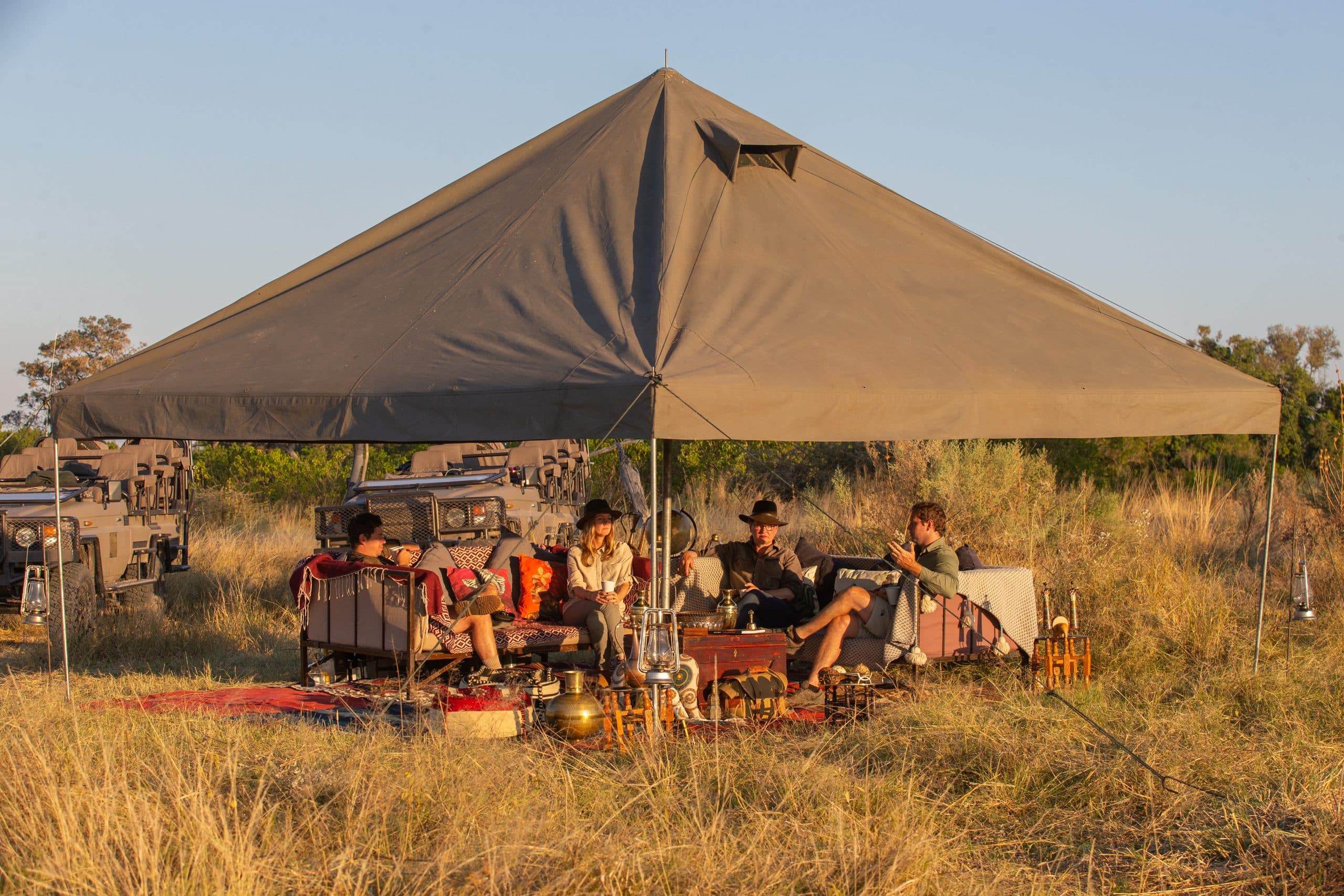 People relaxing under a large beige canopy tent in a dry field, with chairs, tables, and parked safari vehicles nearby at sunset.