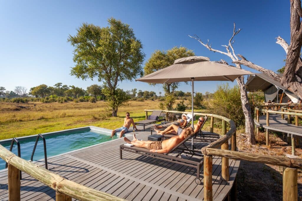 People sunbathing on lounge chairs by a rectangular pool on a wooden deck under a large umbrella in a sunny outdoor setting.