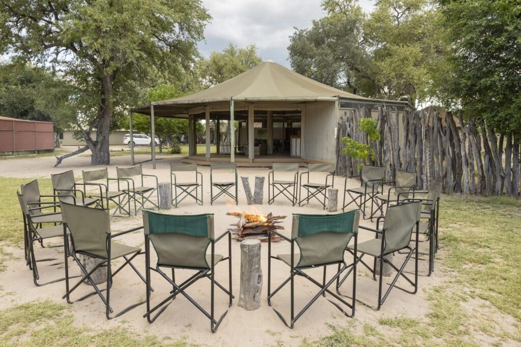 Outdoor campfire circle with gray folding chairs around a small fire pit on sandy ground, under a canopy tent and trees in the background.