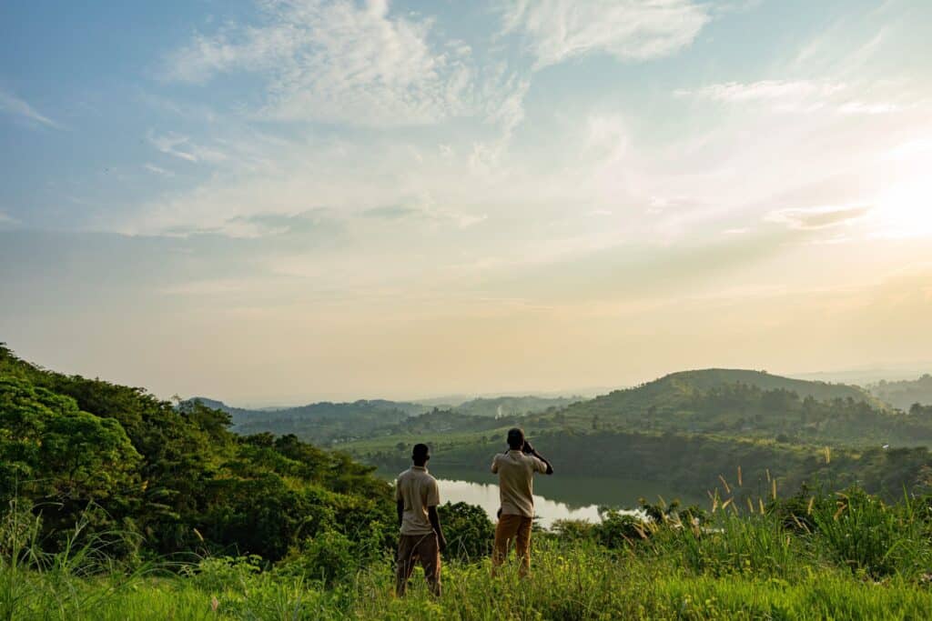 Two people stand on a grassy hill, overlooking a green valley and distant hills under a pale blue sky.