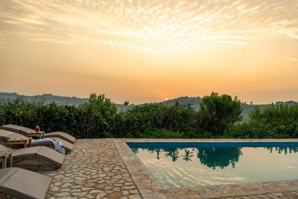 Poolside with stone patio and row of lounge chairs facing a golden sunset over a hilly landscape with green trees in the foreground.