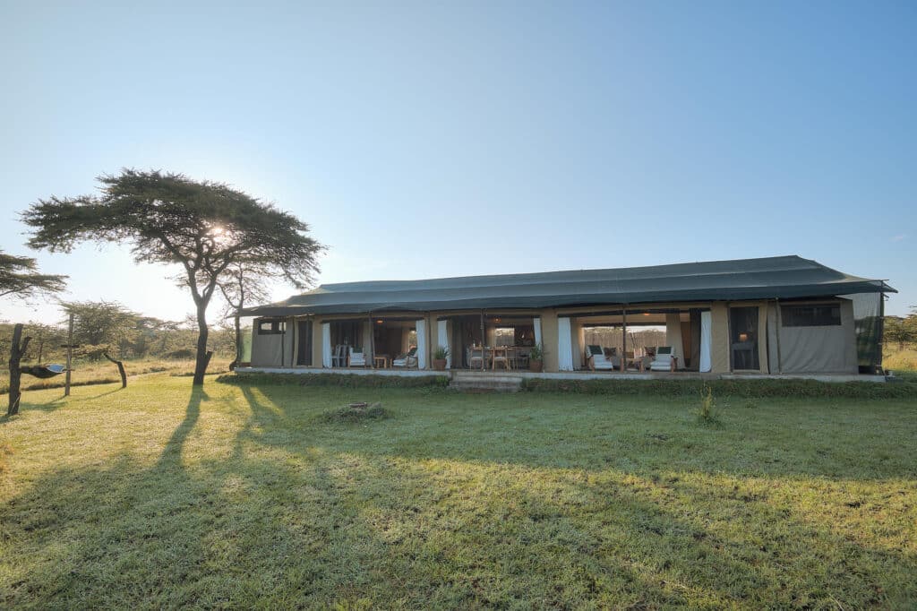 Glamping safari tent with an open veranda and outdoor seating on a sunlit grassy savannah under a clear blue sky