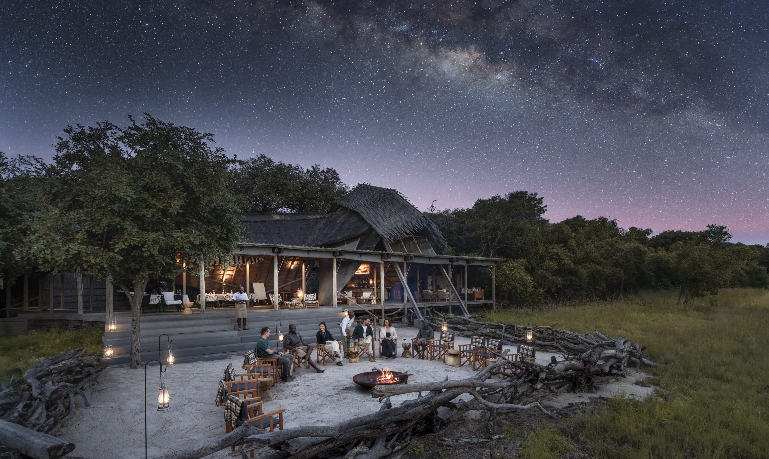 Outdoor dining under a starry night sky with the Milky Way, guests seated around lanterns and a central fire pit.