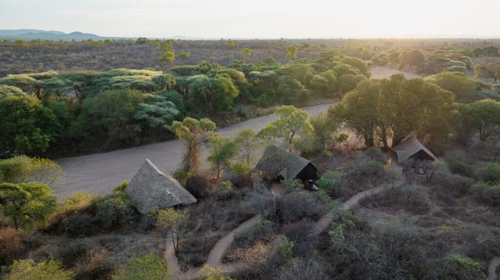 Aerial view of a rural village with thatched huts and a winding dirt road through a green savannah at sunset.
