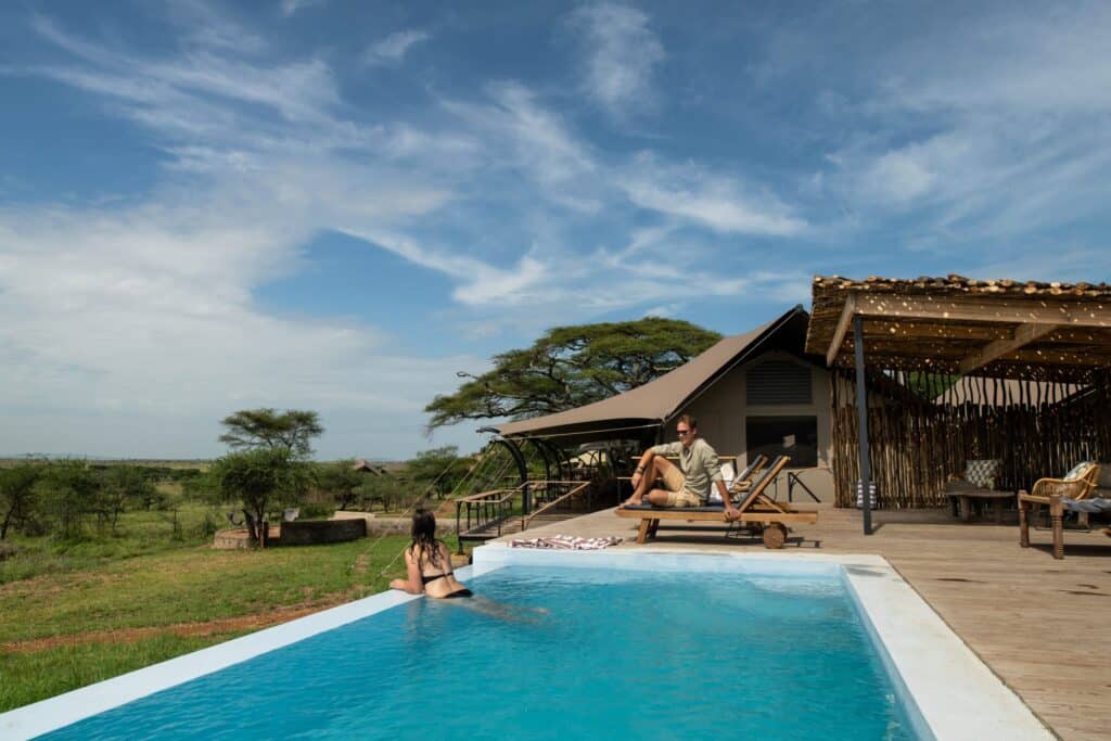 Outdoor pool with a wooden deck at a safari lodge, guests lounging and sunbathing under a thatched canopy on a sunny day with open grassy plains in the background.