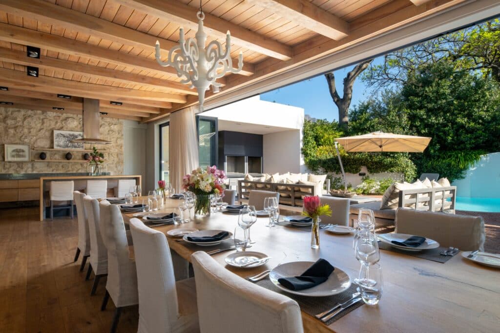 Elegant indoor-outdoor dining area: long table set for a formal meal with glassware, plates, and flower centerpiece under a wooden-beam ceiling and chandelier; pool and patio outside.