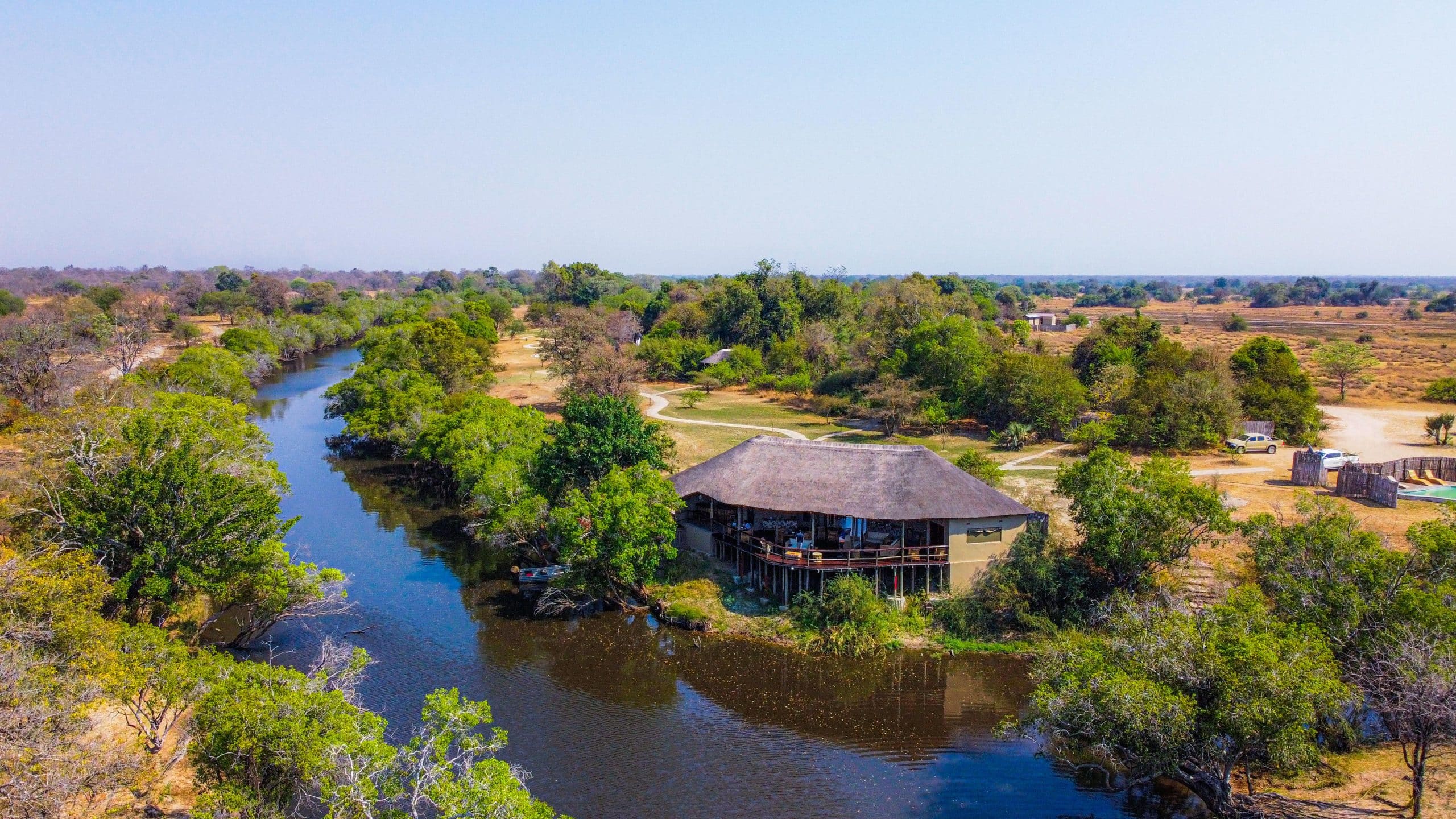Aerial view of a riverside lodge with a thatched-roof building on stilts, surrounded by trees and a winding blue river.