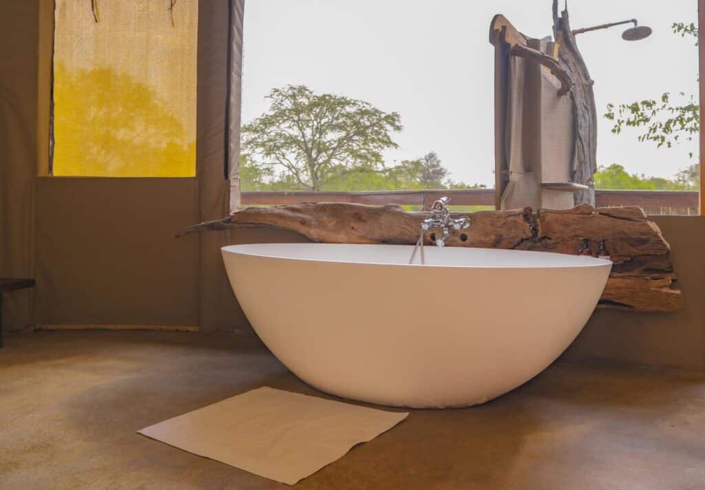 White freestanding bathtub set in a rustic bathroom with a wooden log surround and a window showing trees outside, bright outdoors visible through the window, artful natural feel.