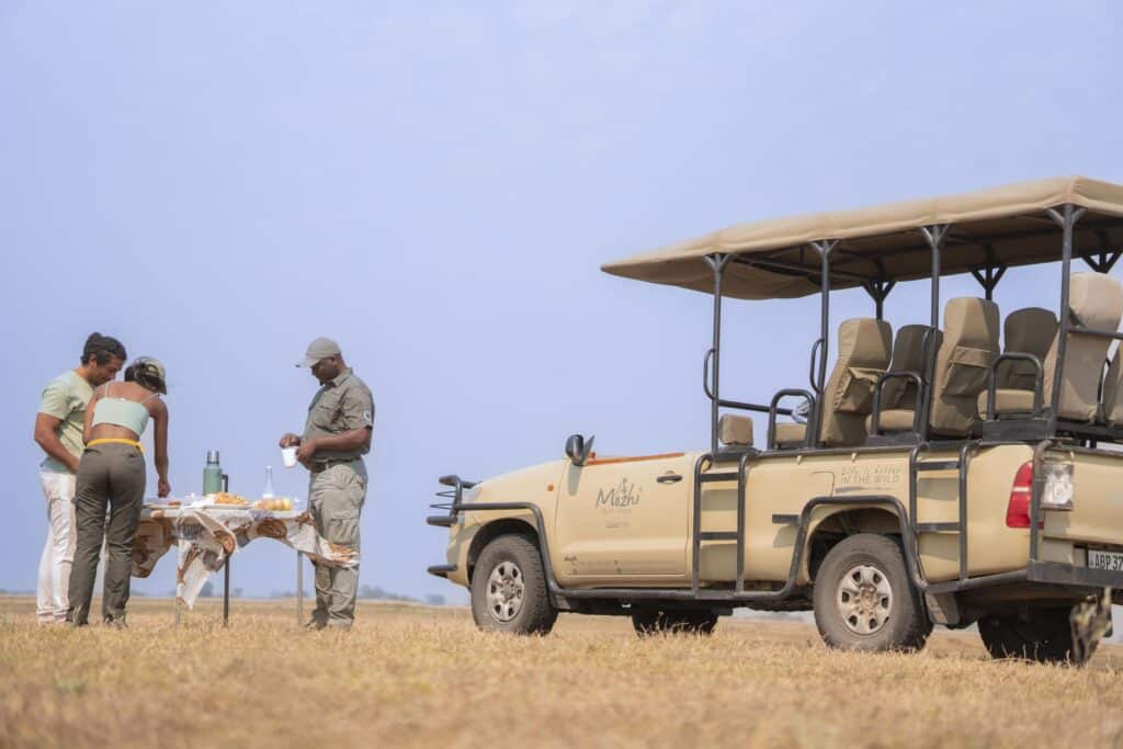 Group of people sharing a meal beside a beige safari jeep with a canopy in a dry, open field. A table with food is set up and one person holds a cup while others chat nearby.