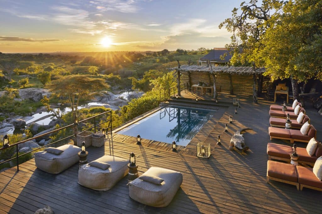 Sunset over a wooden deck by a pool, with lounge chairs and a rustic pergola, overlooking a savannah landscape.