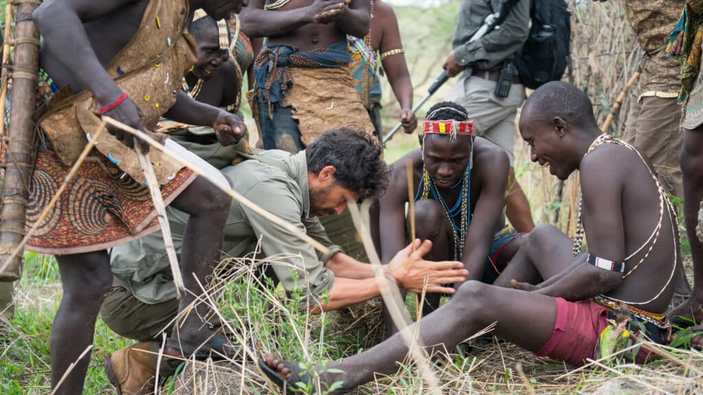 Group of people in traditional dress gathered around a seated man, as others pose with sticks in a rural outdoor setting.