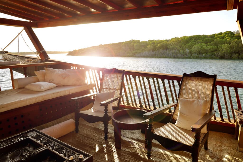 Wooden boat deck with cushioned seating and wicker chairs, overlooking a sunlit river and forested shoreline.