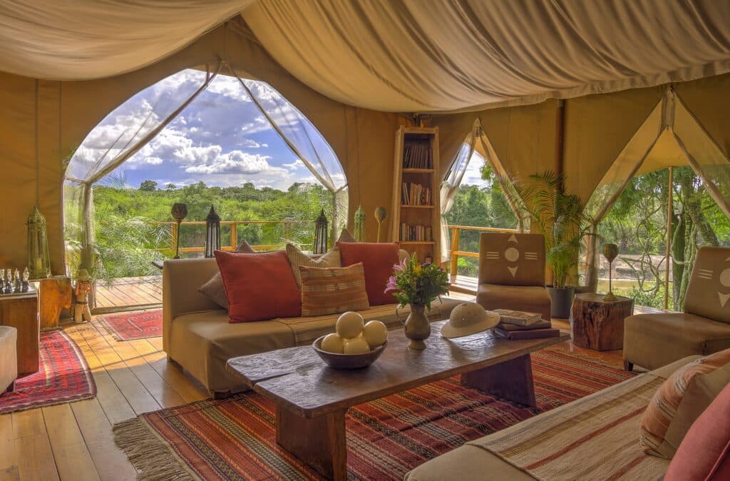 Cozy tent living room with beige sofas, orange pillows, a wooden coffee table, and a view of greenery outside through large openings.