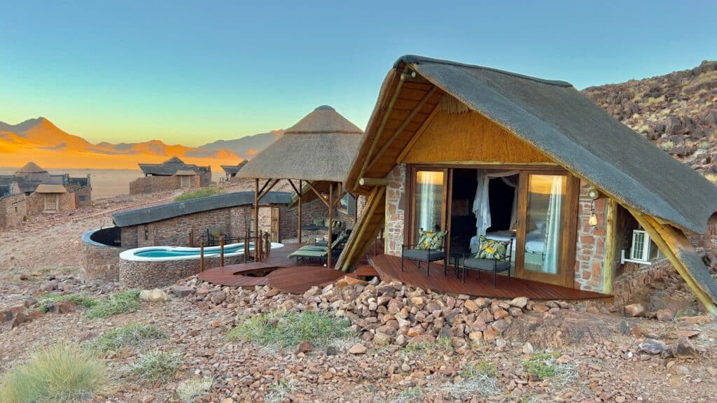 Thatched-roof bungalow with a wooden deck and seating, set in a desert hillside with a stone wall and pool nearby at sunset.