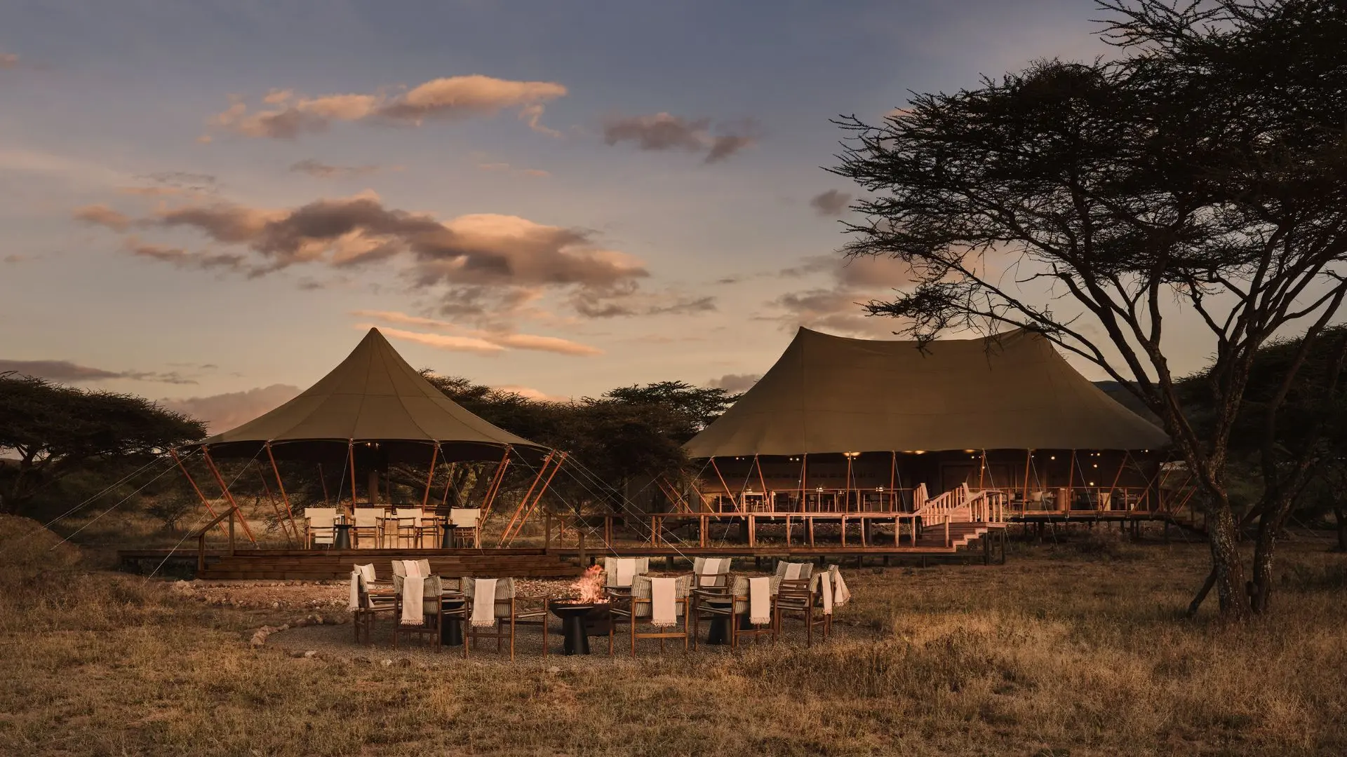 Two large beige canvas tents on a wooden deck with chairs around a fire pit, under a sunset sky with trees nearby.