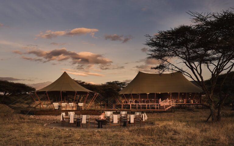 Two large beige canvas tents with tables and chairs set up for an outdoor evening gathering around a central fire pit.
