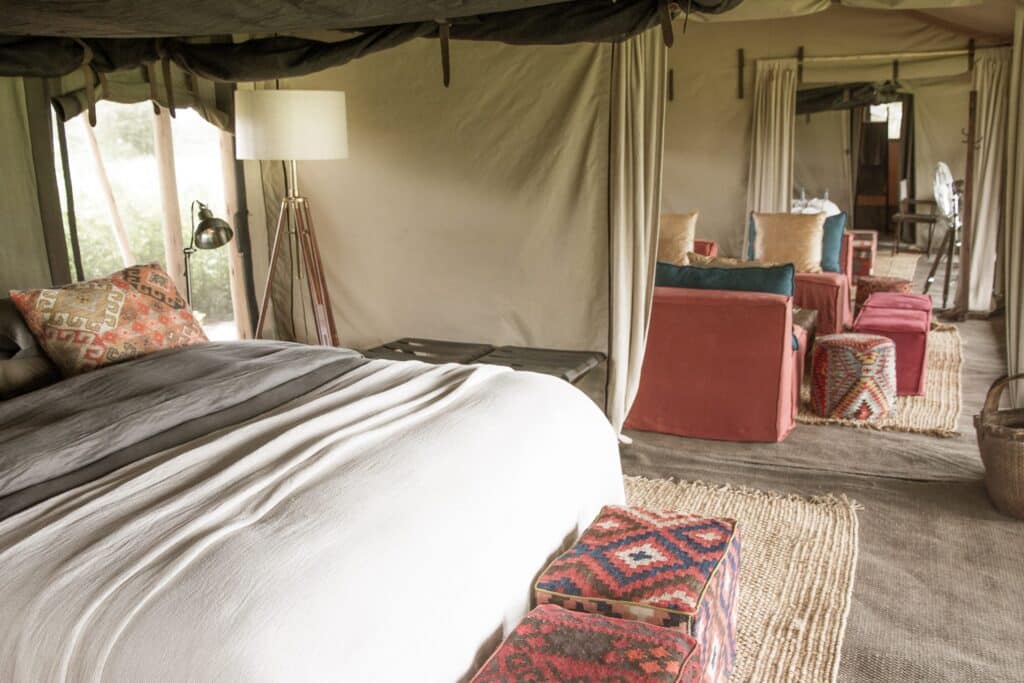 Inside a glamping tent with a large bed in the foreground, beige linens and patterned pillows, and a cozy seating area with colorful cushions farther back.