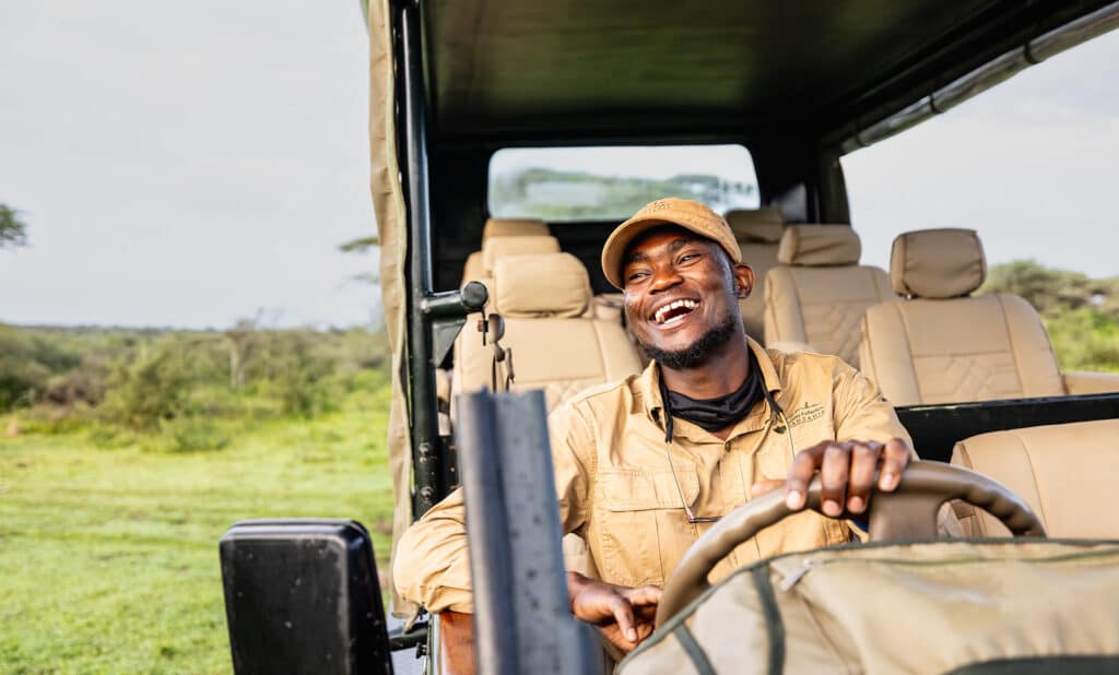 Smiling safari guide in a tan uniform driving an open-top vehicle through grassy savannah.
