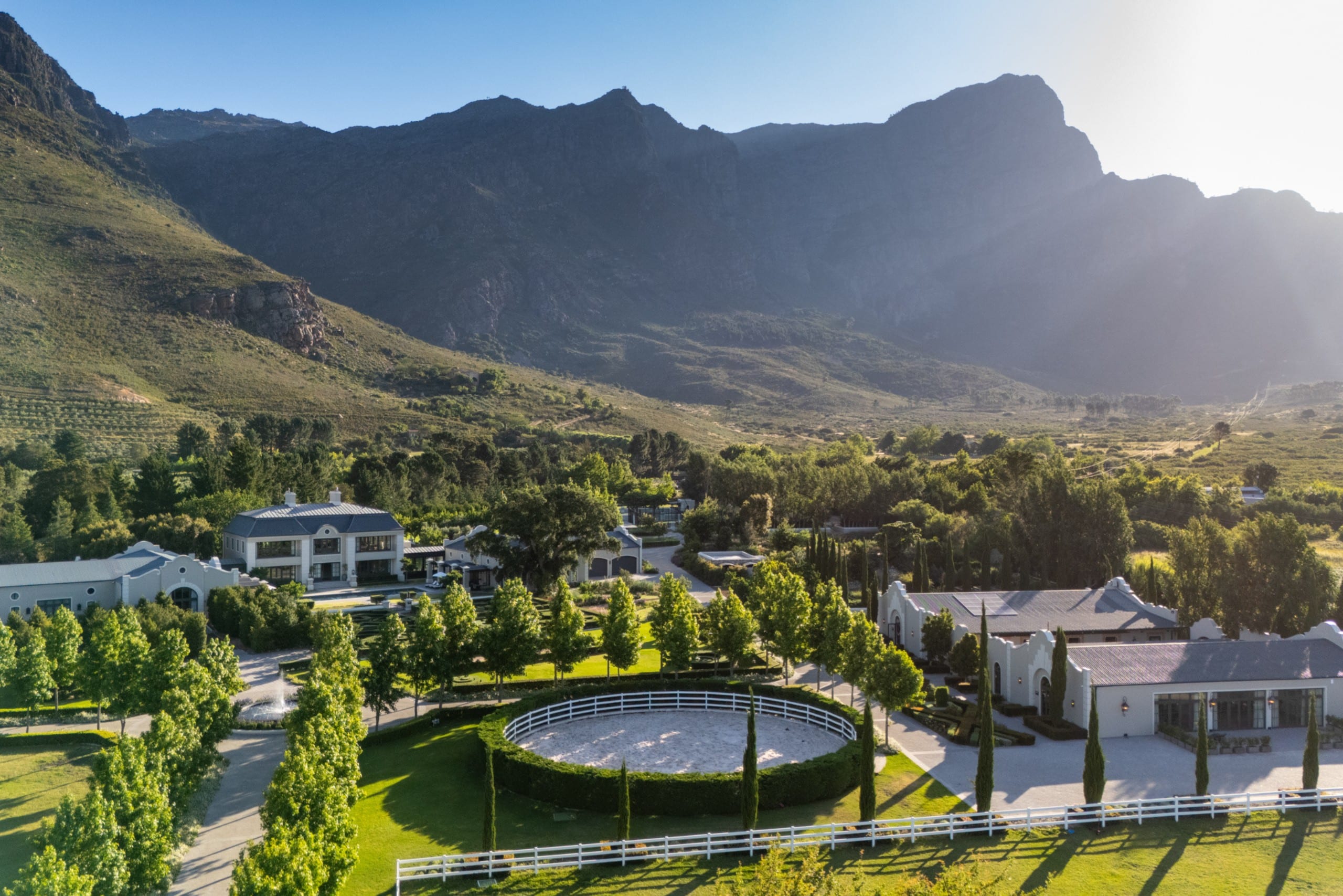Aerial view of a mountain range behind a white estate with manicured lawns and a circular riding arena in the foreground.