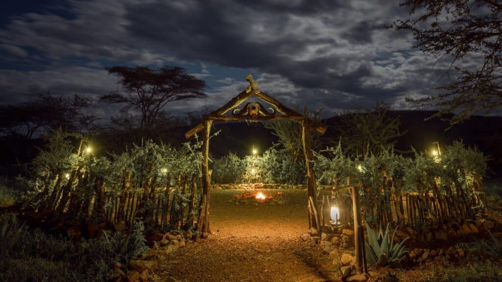 Nighttime garden entrance with a wooden arch, lanterns, and a small fire pit in the foreground.