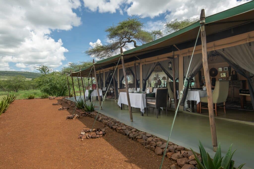 Open-air restaurant under a canvas tent with white-clothed tables, wooden posts, and desert landscaping under a blue sky.