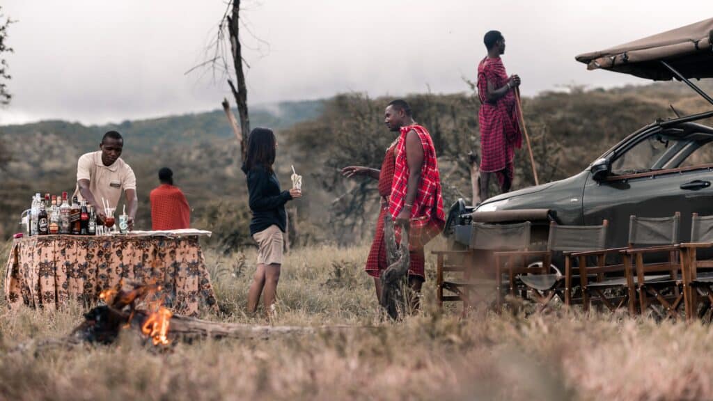 Outdoor camp scene with a table of bottled drinks, a server pouring beverages, and people sharing a drink near a vehicle with a canopy in the background.