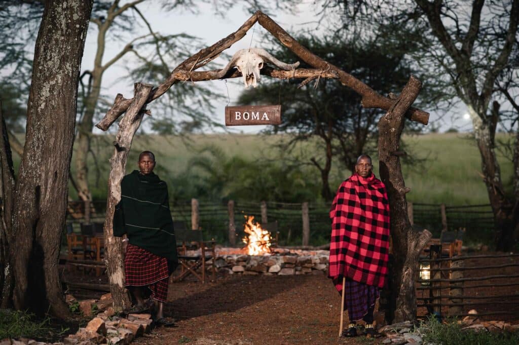 Two people wrapped in red-and-black blankets stand by a campfire under a rustic wooden arch with a hanging animal skull and a 'BOMA' sign.