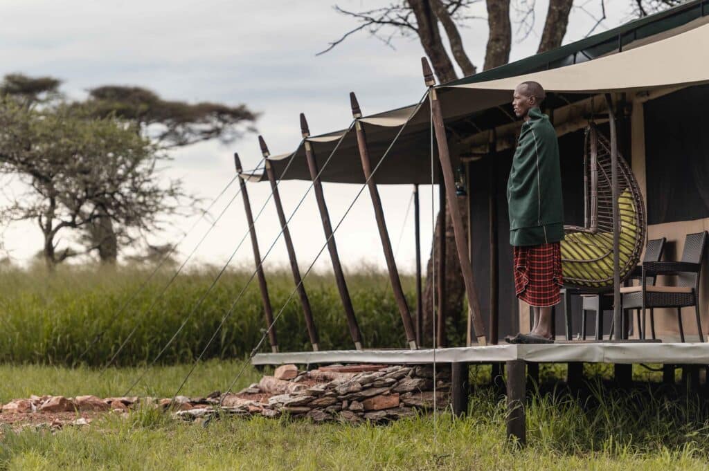 Man stands on a wooden deck beside wicker chairs outside a canvas tent in a savanna setting.