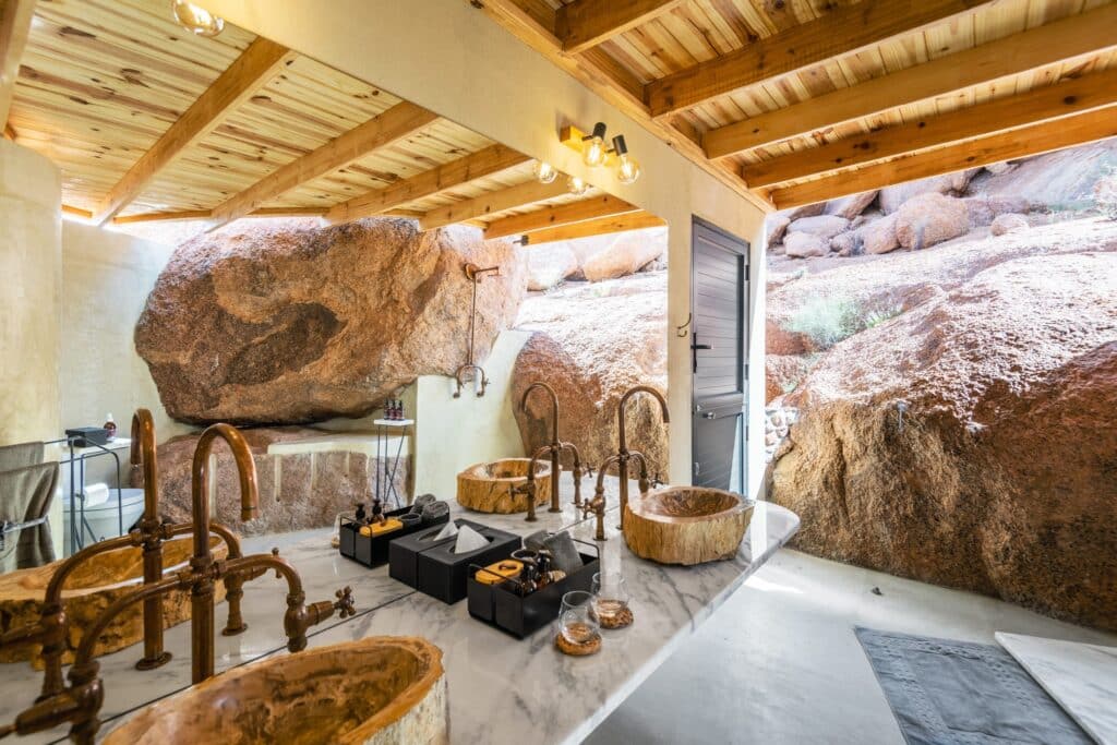 Outdoor rustic bathroom with rock walls, wooden ceiling beams, and copper stone sinks set on a marble counter under warm lighting of exposed bulbs in a rocky setting on a patio.