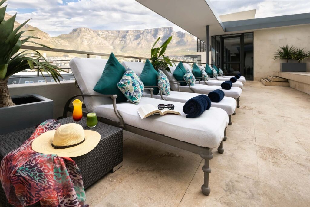 Row of white cushioned lounge chairs with teal and patterned pillows on a rooftop terrace, mountain range in the distance.