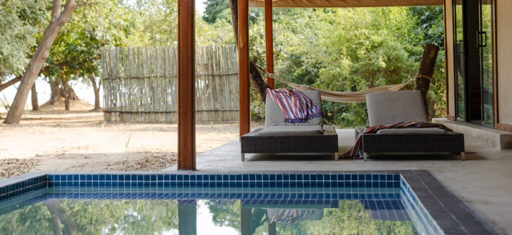 Outdoor patio with wooden posts, beige canopy, and built-in lounge chairs beside a rectangular blue-tiled pool, greenery in the background