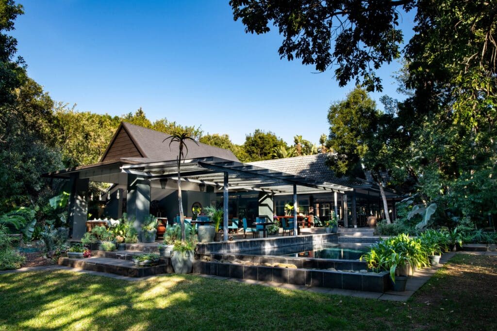 Outdoor patio with dark roofed house, glass doors, and a pergola surrounded by potted plants and greenery beside a stepped pool area.