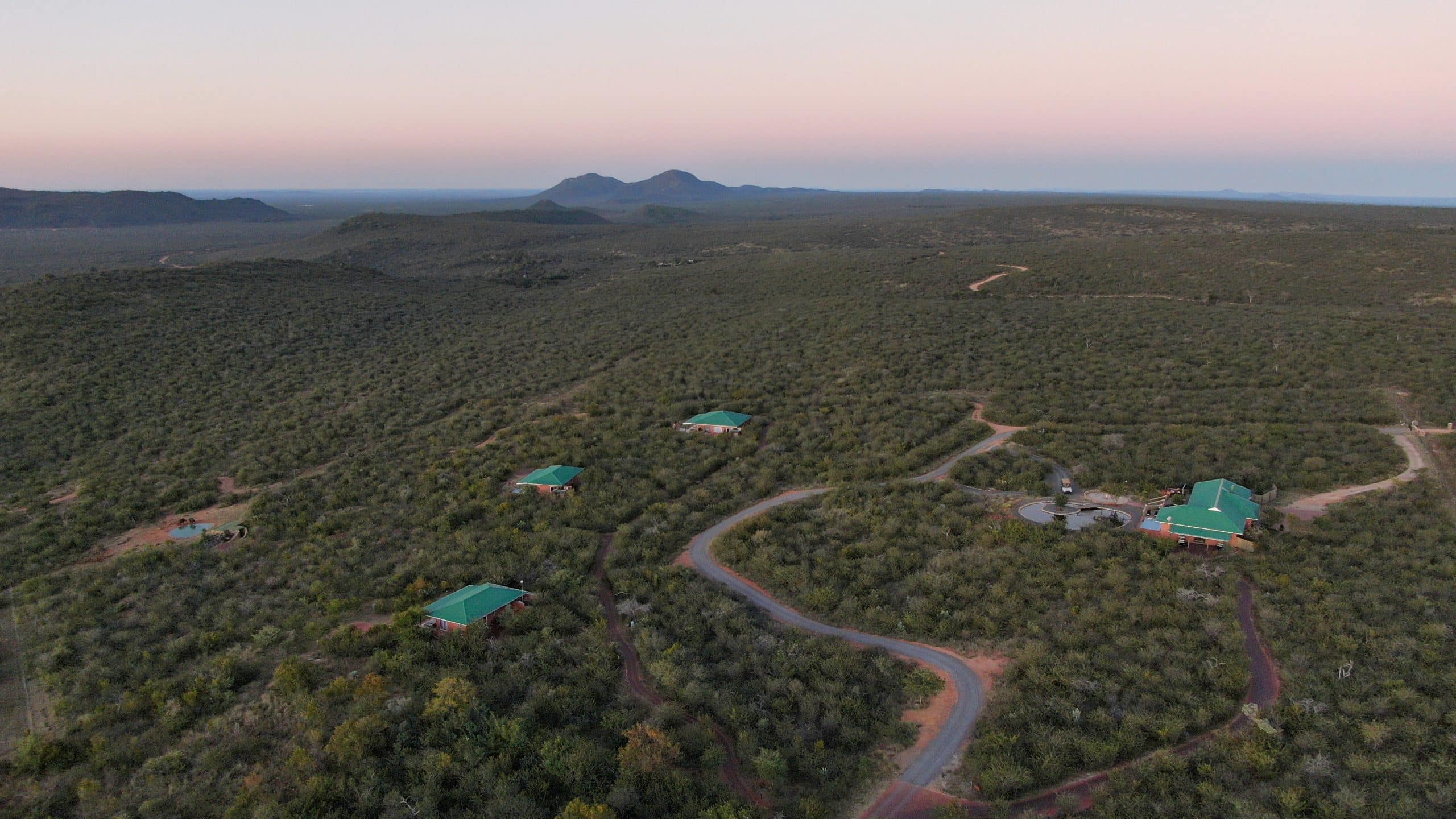 Aerial view of scrubby desert landscape with winding roads and several green-roofed buildings, mountains on the horizon at dusk.