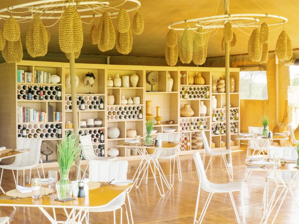 Restaurant interior with hanging woven corn-husk decorations, wooden shelves filled with vases and wine bottles, and white metal chairs.