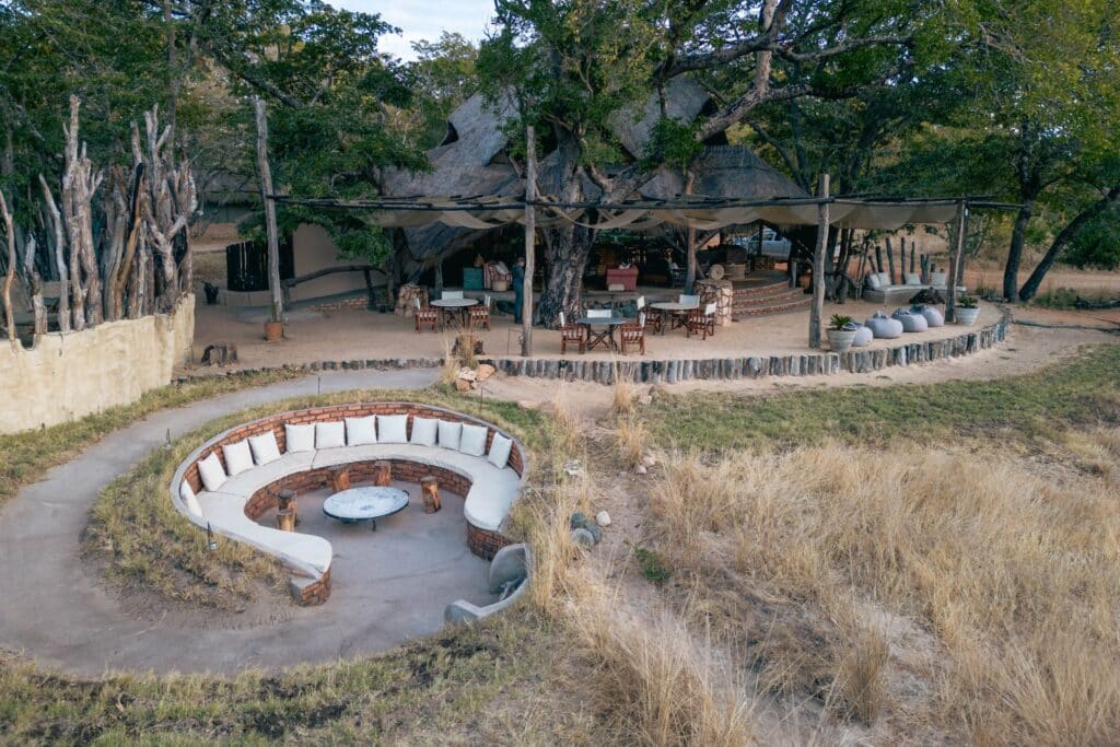 Circular brick seating with white cushions around a central fire pit under a thatched canopy in a wooded outdoor area.