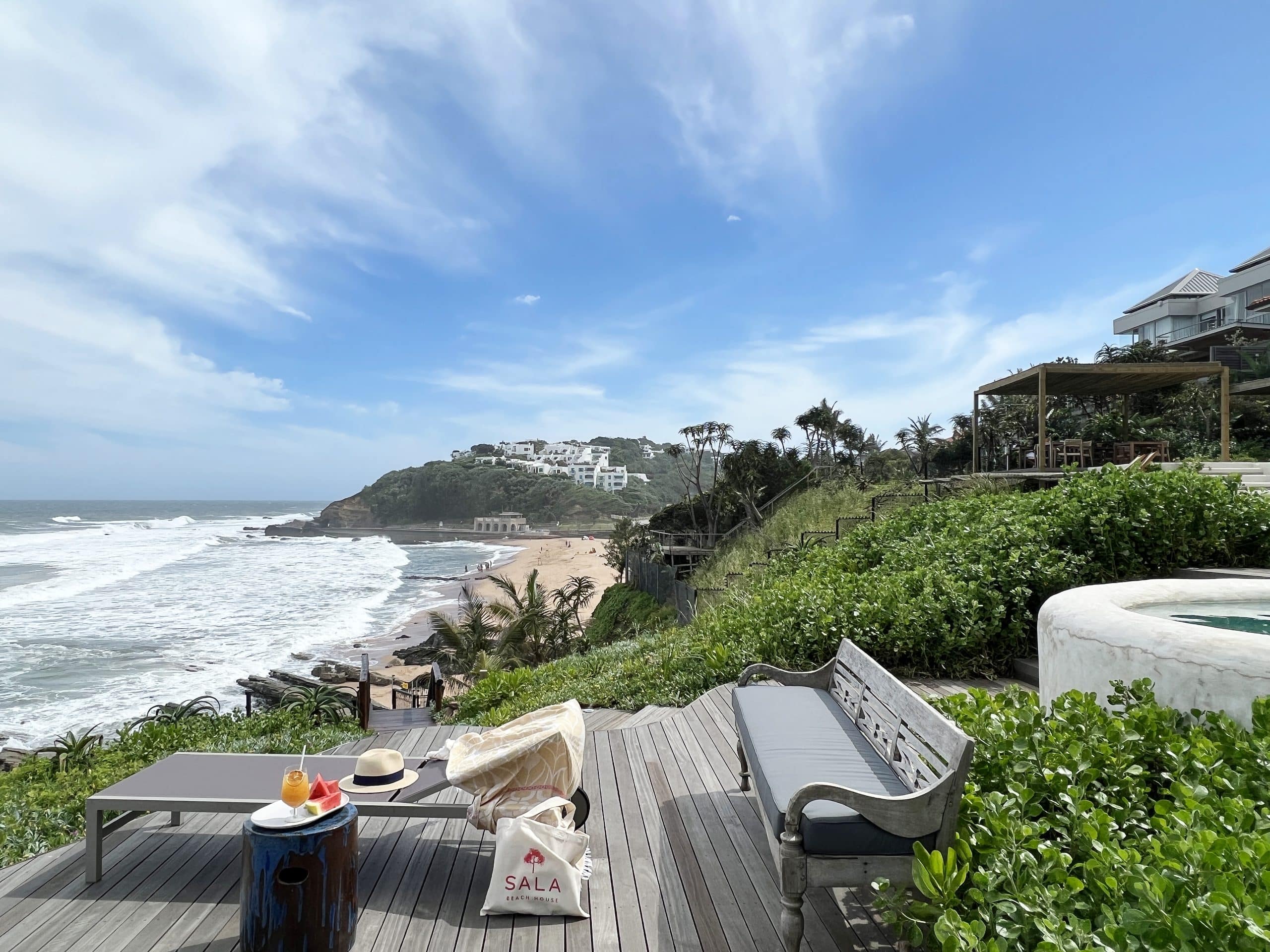 Oceanfront wooden deck with lounge chairs, a table of drinks, hat, and tote bag; view of sandy beach and hillside houses in the distance.