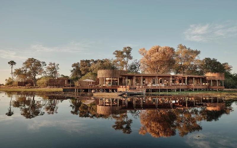 Lakeside wooden lodge with open dining area, trees, and a calm reflection in the water at sunset/early morning.