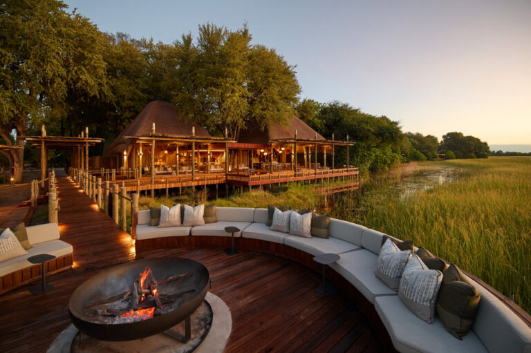 Curved outdoor seating around a lit fire pit on a wooden deck overlooking a marsh at sunset, with thatched-roof huts and trees in the background