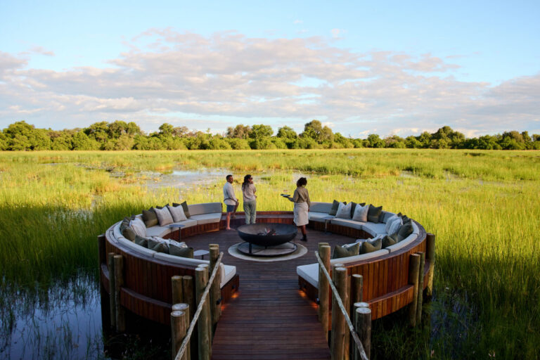 Circular wooden deck with built-in couches surrounding a fire pit, on a marshy landscape with three people chatting under a blue sky.