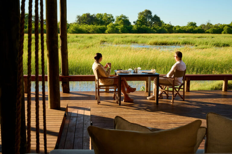 Two people sit at a table on a wooden deck overlooking a grassy wetland and water under a clear blue sky, enjoying a meal.